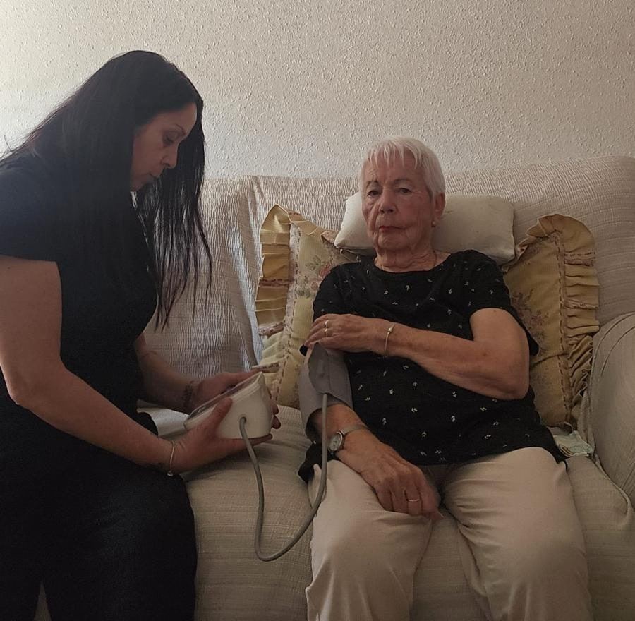 Cuidadora d'Atention prenent la tensió arterial a una senyora gran al sofà de casa seva